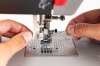 Maszyna do szycia Veritas Power Stitch PRO z widocznym mechanizmem igły, czarną nicią oraz metalową płytką z oznaczeniami do szycia. Ujęcie z bliska, ręka trzymająca nić i elementy maszyny, w jasnym tle.