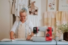 Maszyna do szycia Veritas Power Stitch PRO w kolorze szarym z czerwonymi akcentami, stojąca na niebieskim stole. W tle widoczne materiały do szycia oraz rośliny. Użytkownik, w jasnej koszuli i fartuchu, składa materiał, używając miarki krawieckiej.