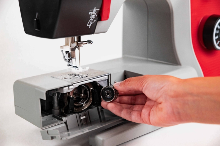 Maszyna do szycia Veritas Power Stitch PRO w kolorze szarym, z widoczną ręką wyjmującą szpulę z bębna. Ujęcie z boku, pokazujące mechanizm szycia oraz panel z 17 ściegami. Obok znajduje się stopka do szycia. Tło białe, co podkreśla detale maszyny.