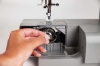Maszyna do szycia Veritas Power Stitch PRO w kolorze szarym, z widocznym mechanizmem chwytacza. Użytkownik trzyma metalowy chwytacz w pobliżu otwartego schowka, ukazując szczegóły konstrukcji. Na górze maszyny widoczne przyciski do wyboru 17 ściegów.