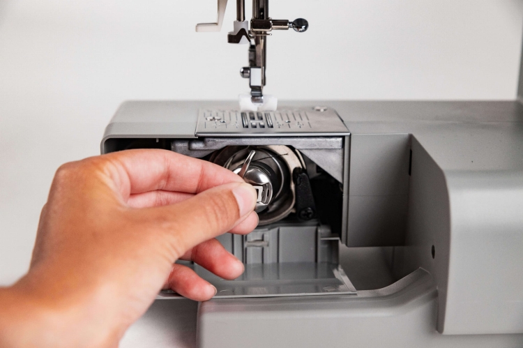 Maszyna do szycia Veritas Power Stitch PRO w kolorze szarym, z widocznym mechanizmem chwytacza. Użytkownik trzyma metalowy chwytacz w pobliżu otwartego schowka, ukazując szczegóły konstrukcji. Na górze maszyny widoczne przyciski do wyboru 17 ściegów.
