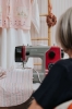 Maszyna do szycia Veritas Power Stitch PRO w kolorze szarym i czerwonym, z 17 ściegami. Widoczna w frontalnym ujęciu, z igłą w pozycji roboczej, szyjąca różowy materiał w paski z wzorem. Tło w jasnych odcieniach, z wiszącymi ubraniami.