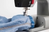 Maszyna do szycia Veritas Power Stitch PRO z widocznym elementem szyjącym, umieszczona na blacie. Na zdjęciu widać fragment denimu z żółtą nicią przeszywającą krawędź materiału. Ujęcie z bliska, pokazujące szczegóły mechanizmu i materiału.