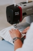 Maszyna do szycia Veritas Power Stitch PRO w kolorze czarnym i szarym, z widocznymi elementami sterującymi. Ujęcie z boku, pokazujące ręce użytkownika pracującego nad białym materiałem z plisami. Tło jasne, proste, skupiające uwagę na maszynie i tkaninie