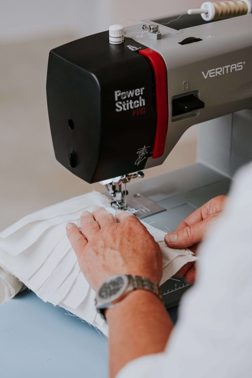 Maszyna do szycia Veritas Power Stitch PRO w kolorze czarnym i szarym, z widocznymi elementami sterującymi. Ujęcie z boku, pokazujące ręce użytkownika pracującego nad białym materiałem z plisami. Tło jasne, proste, skupiające uwagę na maszynie i tkaninie