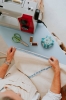 Maszyna do szycia Veritas Power Stitch PRO w kolorze szarym z czerwonymi akcentami, umieszczona na stole. Widać także zielone nożyczki, szpulki z nicią w kolorze brązowym oraz miarkę krawiecką, podczas gdy ręka trzyma materiał.