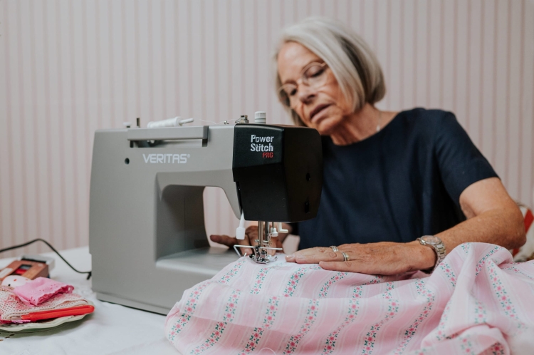 Maszyna do szycia Veritas Power Stitch PRO w kolorze szarym, z 17 ściegami. Ujęcie z boku, z widoczną osobą szyjącą różowy materiał w białe i zielone wzory. Na stole leżą przybory krawieckie. Tło w pasy, jasne i stonowane.