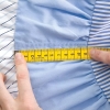 Centymetr krawiecki obustronny o długości 150 cm, wykonany w Niemczech. Miernik leży na tkaninie w kolorze błękitnym, z widoczną skalą pomiarową w odcieniach żółci i czerni, z ręką trzymającą go na końcu.