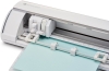 Ploter tnący Silhouette Cameo 5 w kolorze białym, widok z góry. Na urządzeniu umieszczony jest błękitny arkusz papieru, z widocznym mechanizmem tnącym i prowadnicami. Wnętrze ploteru jest dobrze oświetlone, co podkreśla detale konstrukcji.