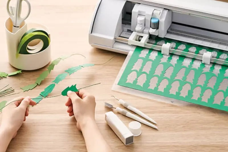 Ploter tnący Silhouette Cameo 5 Plus w kolorze białym z rolką zielonego papieru w procesie cięcia. Na stole widoczne ręce przygotowujące wycięte zielone elementy oraz narzędzia do precyzyjnego wykończenia.