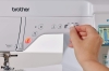 Maszyna do szycia Brother F560 w ujęciu frontalnym. Widoczne przyciski do regulacji, w tym pokrętło do ustawiania szerokości ściegu oraz wyświetlacz LCD. Ręka trzyma pokrętło na białym tle, widać również logo brother.