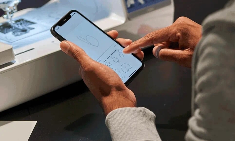 Osoba trzymająca telefon w dłoniach, z widoczną aplikacją do projektowania haftów, na tle maszyny hafciarskiej Brother M380D. Na ekranie telefonu szkice różnych wzorów haftów. Stół w ciemnym kolorze, maszyna w białej obudowie.
