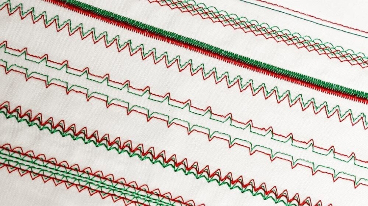 Zbliżenie na fragment tkaniny z różnymi wzorami ściegów, wykonanymi w kolorach czerwonym i zielonym. Widoczne są zygzaki, fale i prostokątne wzory, ukazujące funkcje maszyny do szycia Janome JUNO E1015. Tło białe, podkreślające detale ściegów.