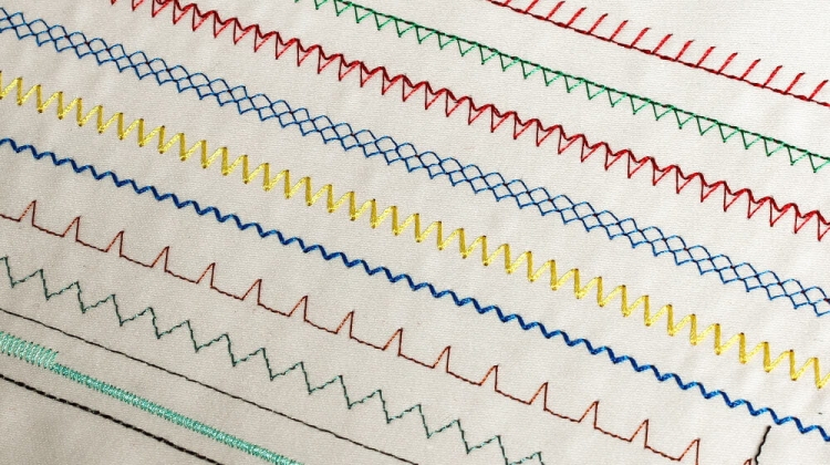 Wzory ściegów wykonanych przez maszynę do szycia Janome JUNO E1015, w kolorach: czerwonym, zielonym, żółtym, niebieskim i czarnym. Ściegi różnią się kształtem, w tym prostymi liniami i zygzakami, na tle jasnej tkaniny.