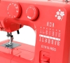 Maszyna do szycia Janome JUNO E1015 w kolorze czerwonym. Widoczne pokrętła do ustawiania ściegów oraz schemat różnych rodzajów ściegów po prawej stronie. Na obudowie biały wzór kwiatowy. Produkt w pozycji frontalnej, zbliżenie na panel sterujący.