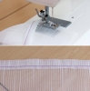 Maszyna do szycia Janome JUNO J15 z widoczną stopką i igłą szyjącą, na jasnym tle. Zbliżenie na materiał z białymi paskami, zszywany na krawędzi z fioletową nitką. W tle drewniana powierzchnia robocza.