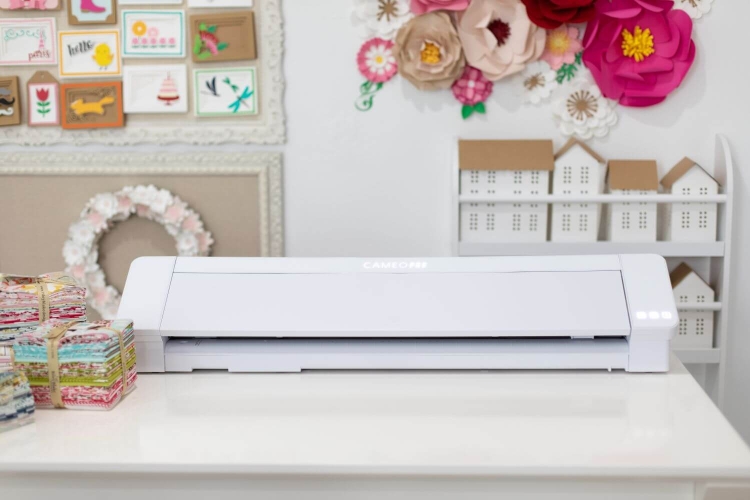 Ploter Silhouette Cameo 4 Pro w kolorze białym, o prostokątnym kształcie, umieszczony na jasnym blacie. Widoczne oświetlenie LED z przodu. W tle kolorowe arkusze papieru oraz dekoracyjne elementy.