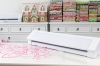 Ploter Silhouette Cameo 4 Pro w kolorze białym, leżący poziomo na stole, z widoczną różową grafiką cięcia. Tło z kolorowymi materiałami i ozdobnym domkiem z piernika. Urządzenie przeznaczone do precyzyjnego cięcia materiałów do 60 cm.