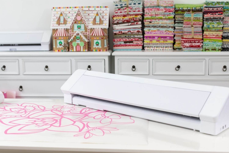 Ploter Silhouette Cameo 4 Pro w kolorze białym, leżący poziomo na stole, z widoczną różową grafiką cięcia. Tło z kolorowymi materiałami i ozdobnym domkiem z piernika. Urządzenie przeznaczone do precyzyjnego cięcia materiałów do 60 cm.