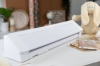 Ploter Silhouette Cameo 4 Pro w kolorze białym, o prostokątnym kształcie, z otwartą przestrzenią tnącą. Ujęcie z boku, ukazujące detale obudowy i mechanizmu. W tle widoczne resztki materiałów oraz dekoracje, tworzące kreatywną atmosferę.