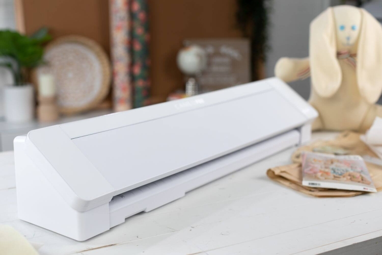 Ploter Silhouette Cameo 4 Pro w kolorze białym, o prostokątnym kształcie, z otwartą przestrzenią tnącą. Ujęcie z boku, ukazujące detale obudowy i mechanizmu. W tle widoczne resztki materiałów oraz dekoracje, tworzące kreatywną atmosferę.