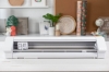 Ploter Silhouette Cameo 4 Pro w kolorze białym, stojący, z widocznym otwartym mechanizmem tnącym. Elementy sterujące umieszczone z przodu. Tło w jasnych kolorach z dekoracjami, jak rośliny i rolki papieru.