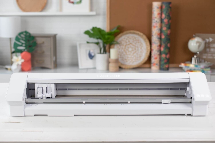 Ploter Silhouette Cameo 4 Pro w kolorze białym, stojący, z widocznym otwartym mechanizmem tnącym. Elementy sterujące umieszczone z przodu. Tło w jasnych kolorach z dekoracjami, jak rośliny i rolki papieru.