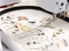 Program Bernina V9 Creator Full Version do projektowania haftów, przedstawiający haftowane wzory z kwiatami, pszczołą i dekoracyjnymi elementami na białym tle. Widoczna maszyna do haftu z igłą w ruchu, w bliskim ujęciu.