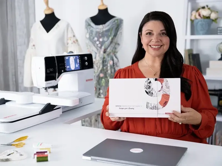 Program Bernina V9 Creator w pudełku, trzymany przez kobietę w czerwonej bluzce. W tle maszyna do szycia Bernina B770 oraz manekiny z haftowanymi sukienkami. Na stole laptop i przybory krawieckie.