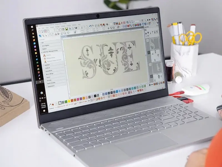 Program Bernina V9 Creator wyświetla projekt haftu na laptopie. Na ekranie widoczny jest napis SUE z dekoracyjnymi wzorami. Laptop jest umieszczony na biurku obok przyborów do szycia, takich jak igły i klej.