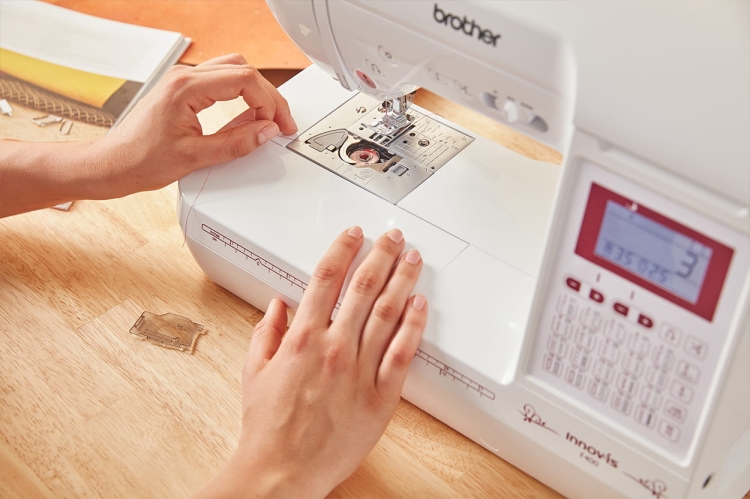 Maszyna do szycia Brother F400 w kolorze białym, z wyświetlaczem LCD i panelem sterującym. Zbliżenie na rękę użytkownika, która nawleka nić przez igłę. W tle widoczne akcesoria do szycia na drewnianym blacie.