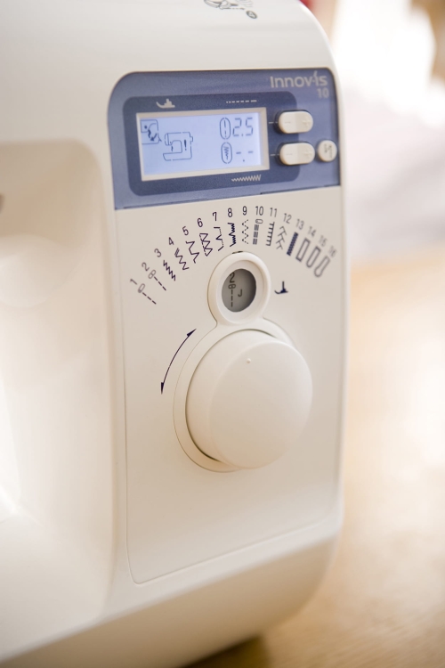 Maszyna do szycia Brother NV15 w kolorze białym, z wyświetlaczem LCD i pokrętłem do wyboru ściegów. Widoczne różne wzory ściegów oraz oznaczenia. Ujęcie z boku, pokazujące detale panelu sterującego.