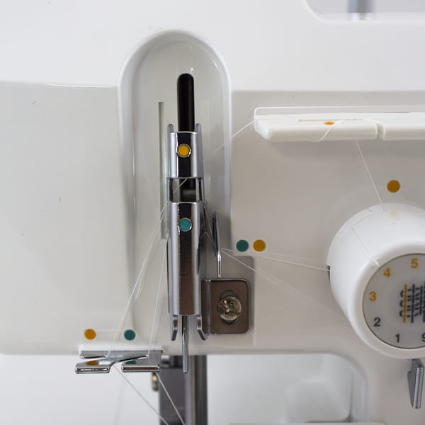 Zbliżenie na mechanizm owerloka JUKI MO-644D, pokazujący igłę, prowadnik nitki oraz pokrętło ustawień. Widoczne kolory oznaczeń ułatwiających nawlekanie nitek. Maszyna w kolorze białym, z metalowymi elementami.