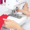 Owerlok TEXI JOYLOCK 2/4 4-nitkowy w białej obudowie, z widoczną igłą i górnym chwytaczem, obszywa czerwony materiał w białe kropki. Ręka operatora trzyma kawałek tkaniny blisko maszyny, na której widoczne są oznaczenia nitkowania.